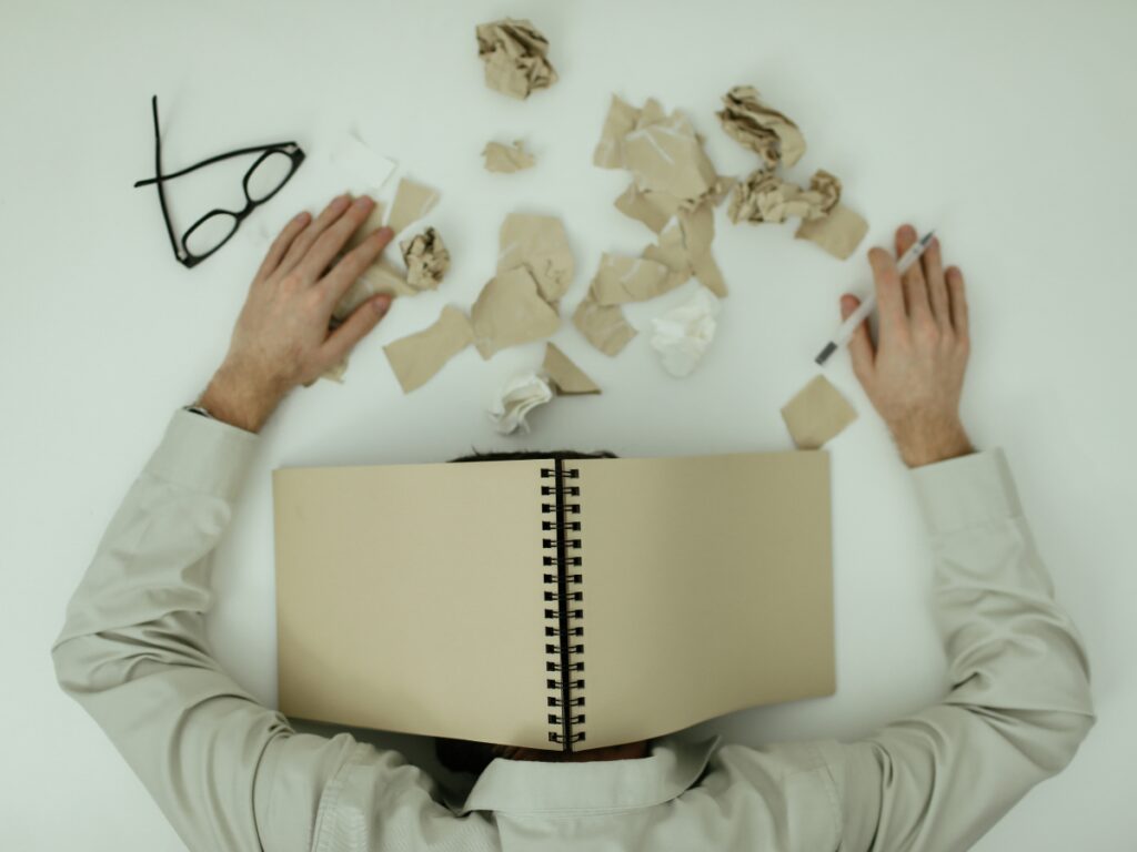 Exhausted person buried under a heavy book with glasses and crumpled paper strewn around.